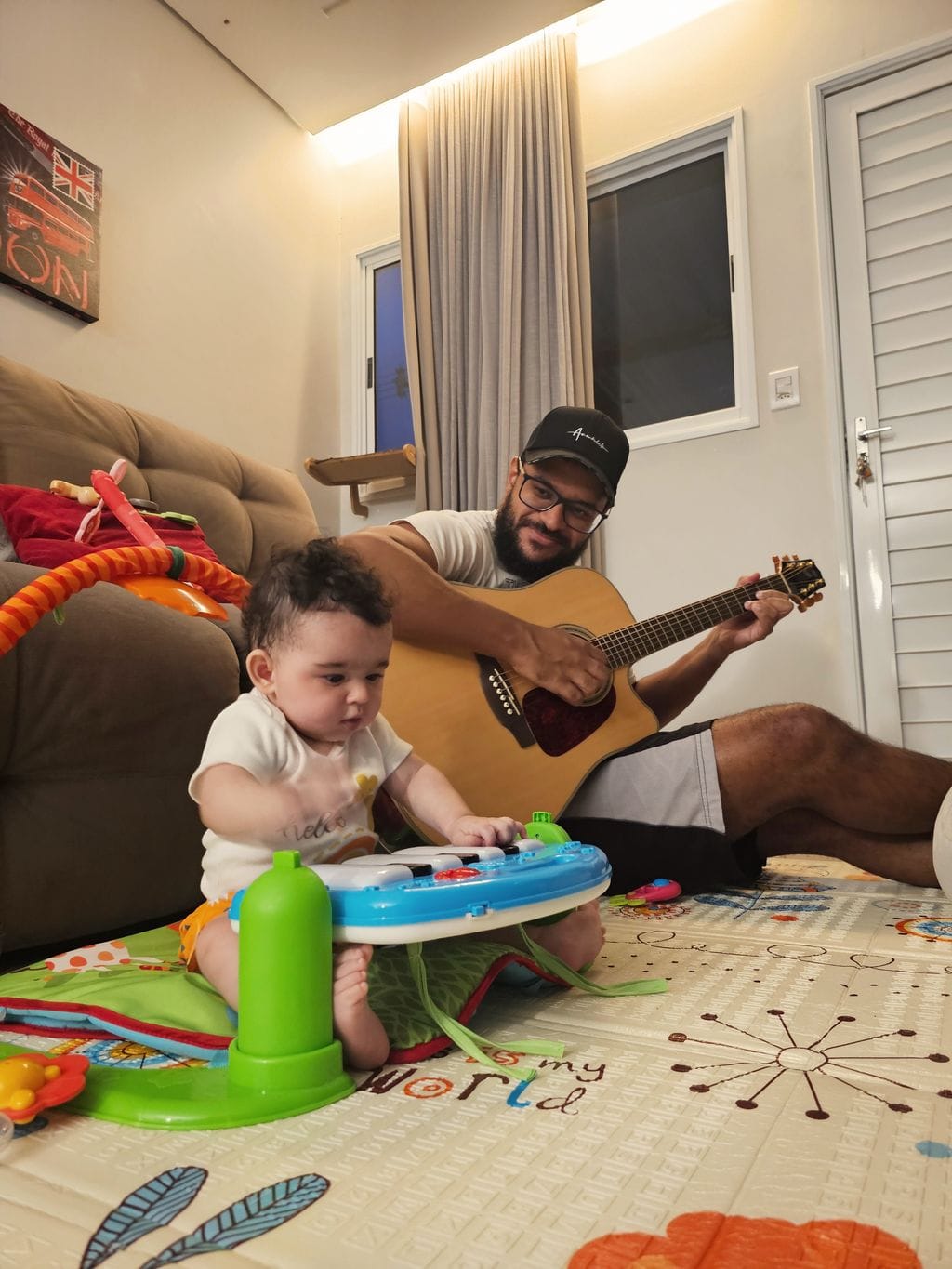 Claudemir tocando violão de pernas cruzadas no tapete de brinquedo com sua filha pequena ao lado tocando um piano infantil